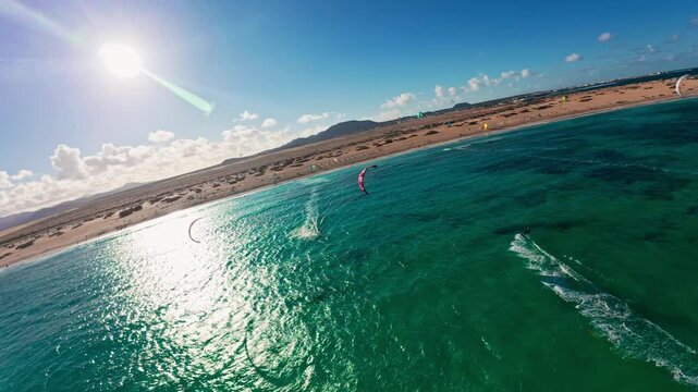 Aerial drone view shows turquoise Atlantic, Corralejo dunes, and volcanic ridges in Fuerteventura. Kite surfers streak across sunlit water with white wake trails.