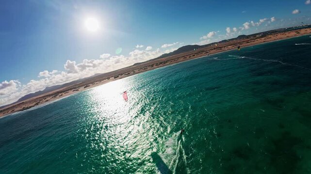 Aerial view shows Fuerteventura coastline in Canary Islands, wide sandy dunes and low volcanic hills, vivid turquoise water, kite surfers, and a circular wake at midday.