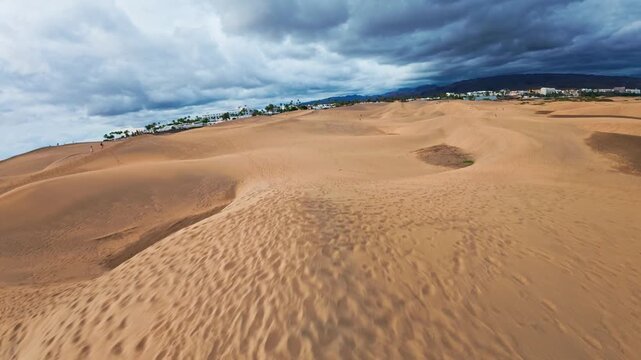 Aerial sweep over Maspalomas Dunes, Gran Canaria. Drone travels along dune ridgelines, footprints and tiny figures visible, palm trees, white resorts, distant mountains.