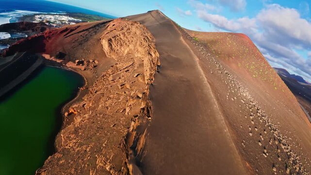 Aerial camera glides over conical volcanoes, lava rims, coastal road, and the emerald lake at El Golfo in Lanzarote, revealing cliffs and a small white settlement.