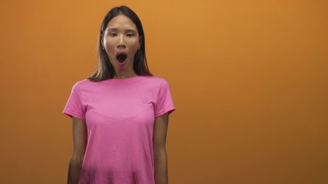 Thai woman with mouth open yawning in studio wearing pink t shirt against orange backdrop, frontal standing pose and visible earrings; fatigue boredom rest.