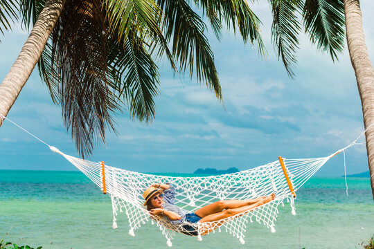 Happy beautiful girl in a straw hat and shorts, striped t-shirt, lying on a beach hammock between two palm trees, on the seashore of a tropical island, traveling summer vacation