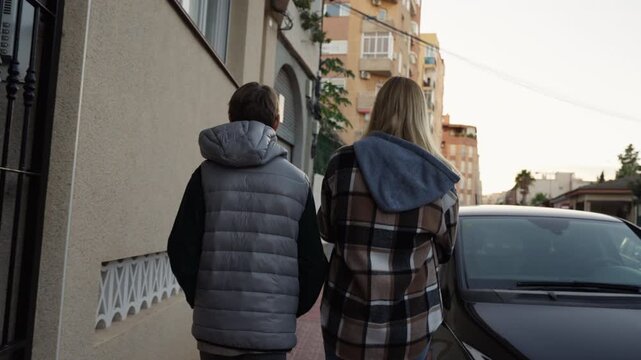 Rear view of mother and son walking together along sidewalk in an urban neighborhood, representing family bonding, communication between parent and teenager, and everyday life