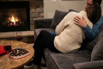 A rear view of a romantic couple embracing on a teal velvet armchair in front of a warm fireplace, accompanied by glasses of red wine and a heart-shaped box of chocolates on a wooden side table.