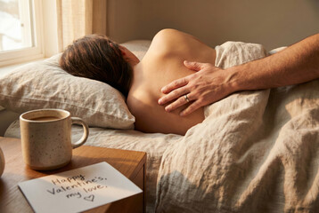 A gentle hand wearing a wedding ring touches the bare shoulder of a sleeping partner in a sunlit bedroom, accompanied by a cup of coffee and a handwritten "Happy Valentine's" note on the bedside table