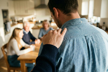A rear view of a man in a denim shirt receiving a comforting hand on his shoulder, with a concerned family group sitting at a kitchen table in the blurred background.