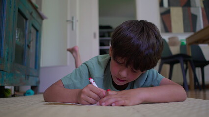 Fototapeta premium Boy lies on floor concentrating while drawing with red marker, indoor environment with furniture and patterned artwork in background