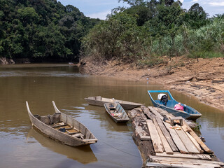 Traditional wooden longboats moored at a rustic wooden jetty on a muddy tropical riverbank surrounded by dense rainforest.