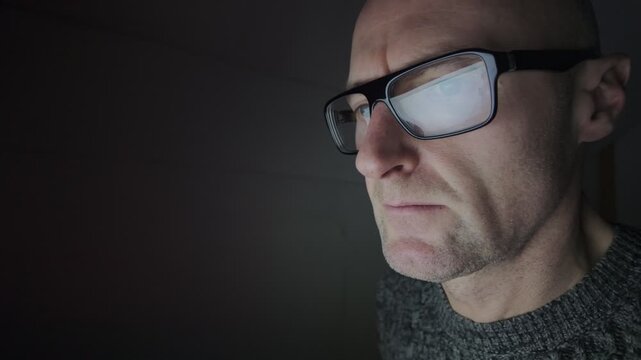 Man sat in dark room using a computer, refection in glasses. Hacker.