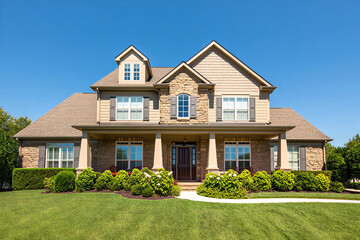 Beautiful large beige house with stone facade and brown roof and door on green lawn with clear blue sky