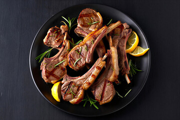 Grilled lamb chops with rosemary and lemon slices on a black plate on dark background