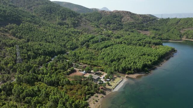 Aerial drone view of a small campsite and RV parking area on the forested shores of Lake Komani, Albania, featuring turquoise water and rugged mountain peaks in the Balkan wilderness.