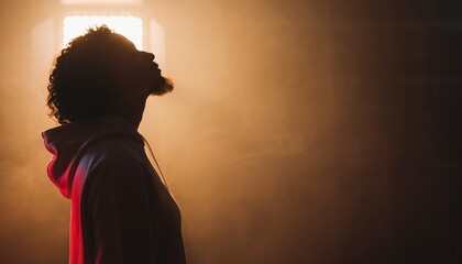 Profile silhouette of a young man with curly hair looking up with hope, illuminated by a warm golden backlight in a dark, smoky room with atmospheric haze and copy space