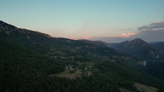 Aerial view of a rafting campsite nestled in the lush Tara River Canyon, Montenegro, with the historic Durdevica Tara Bridge visible in the distance between the steep, forested mountain peaks.