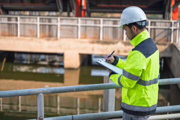 Professional Asian engineer inspecting water quality at a wastewater treatment plant. He is carefully recording maintenance data on a clipboard during an industrial site safety survey.