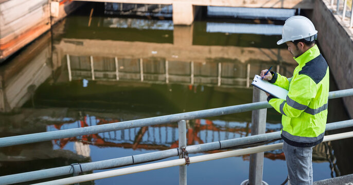 Professional Asian engineer inspecting water quality at a wastewater treatment plant. He is carefully recording maintenance data on a clipboard during an industrial site safety survey.