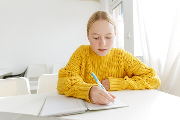 Teenage girl writing in notebook at white table, focused student studying at home, education concept, learning process, school homework