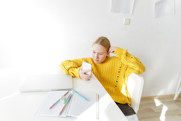Teenage girl sitting at desk with smartphone and school supplies, distracted student during home study, modern education, digital distraction, copy space, bright minimalist interior