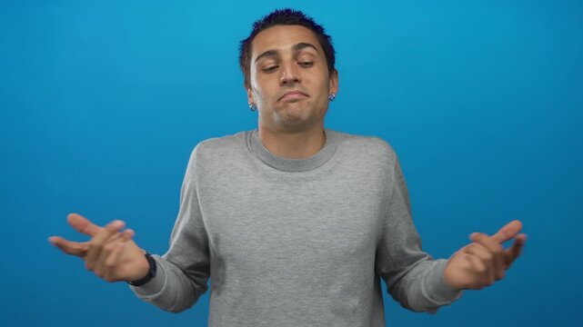 Young hispanic man shrugs with uncertain expression against isolated blue background wearing grey sweater