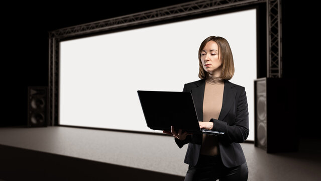 Business presentation and event technology. Professional woman uses laptop on stage in front of large blank screen, preparing digital presentation, conference content, or multimedia display.