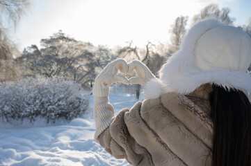 Woman in fur coat making heart shape with hands in snowy park, winter love concept