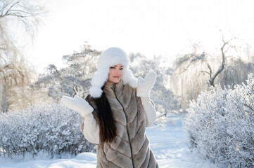 Beautiful woman in white fur hat posing in snowy winter forest during golden hour light
