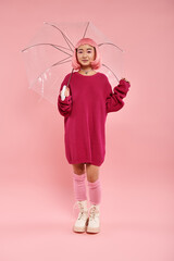 Joyful young woman in pink sweater with clear umbrella against a pink backdrop