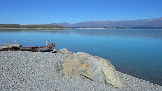 Klarer, ruhiger See mit Ufer und Treibholz vor einer fernen Bergkette unter blauem Himmel, Lake Pukaki Viewpoint, Pukaki, Canterbury, S&uuml;dinsel, Neuseeland, Ozeanien