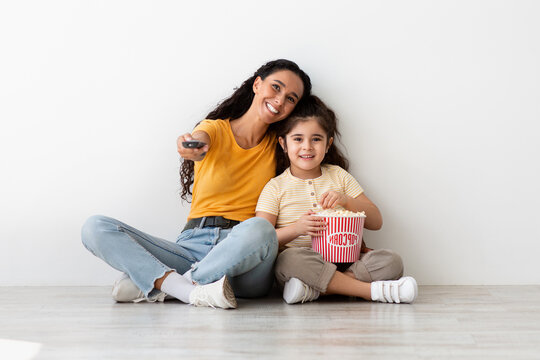Mother Daughter Leisure. Happy Young Woman And Her Female Child Watching Tv Together, Cheerful Little Kid And Mom Holding Remote Controller And Eating Popcorn While Sitting On Floor, Free Space