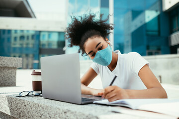African American Girl In Face Mask Learning Online At Laptop And Writing Taking Notes In Notebook During Distance Virtual Lecture Sitting In Urban Area Of Modern University Campus. E-Learning