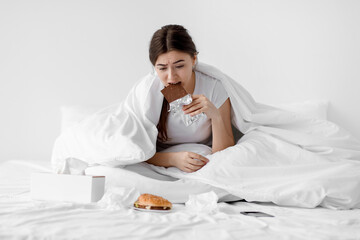 Sad hungry european millennial female eating big chocolate bar sitting on white bed with fast food in bedroom interior. Overeating, meal addiction, stress, loneliness and substitution, copy space