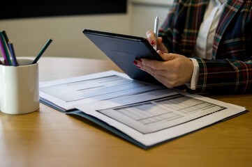 Fototapeta premium A woman in a colorful plaid blazer works on a digital tablet with a stylus, with financial spreadsheets and documents on the desk.