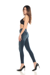 Fototapeta premium Side view full body studio shot of a young woman in black bra and jeans standing in profile on a white background