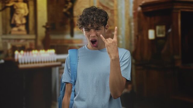 Teenage boy wearing blue backpack flashes rock hand gesture and sticks tongue visible in front of votive candles inside ornate building; playful irreverence.