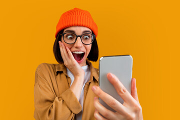 Omg. Surprised hipster woman looking at smartphone screen with shocked face expression, emotionally reacting to good news, standing with phone on yellow background