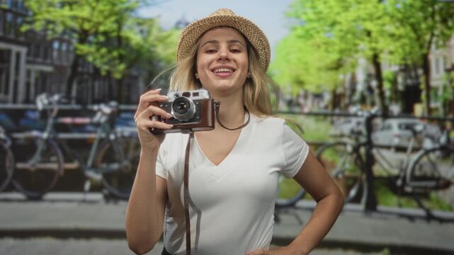 Woman holding camera on street bridge in amsterdam, puffed cheeks and straw hat; playful exploration.