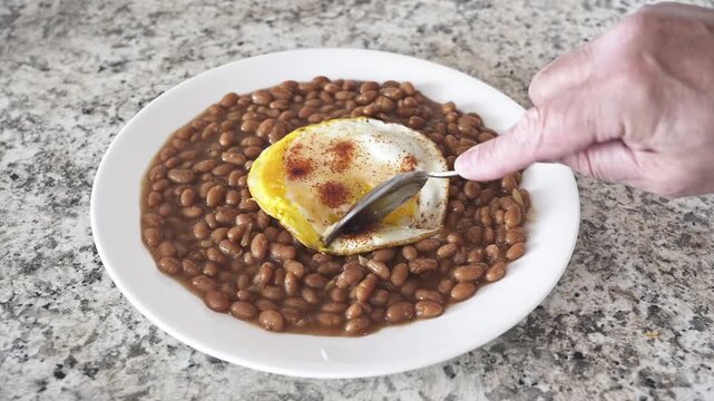 Person eating a fried egg with a runny yolk served over brown beans for a protein-rich breakfast
