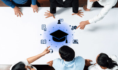 A diverse group of professionals interacts around a table featuring a graduation cap icon, symbolizing education and teamwork in a modern office environment. Trope