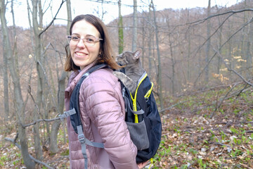 Happy woman smiling and hiking with her gray cat in the backpack in the forest outdoors in spring © DaliCeMedia