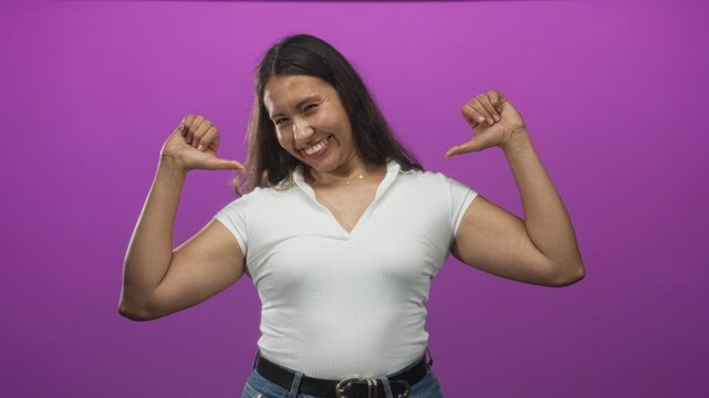 Young hispanic woman pointing thumbs to chest in purple studio wearing white shirt and jeans; self confidence.