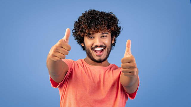 A young man smiles broadly while giving two thumbs up. He wears a pink shirt and poses against a blue background. His happy expression shows excitement and positivity in the moment.