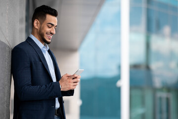 Fototapeta premium Handsome middle eastern businessman in suit standing by modern business center, using cellphone and smiling, sending emails while having break, panorama with copy space, side view