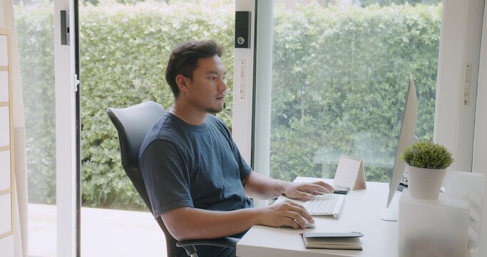 Asian man working intently on desktop computer with keyboard and mouse in bright home office by a window with lush greenery outside, representing focused productivity and modern remote work.