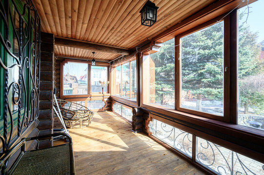Wooden veranda with large panoramic windows, log ceiling, wicker chair, decorative wrought iron railings, lantern lighting &mdash; bright, airy, cozy space with garden views.