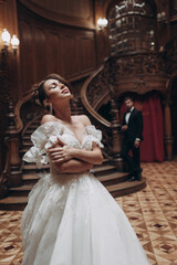 Fototapeta premium Bride in white dress poses on staircase in old mansion with guest in background