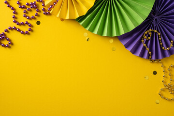 Obraz premium A bright yellow backdrop showcases green and purple paper fans, gold beads, and scattered confetti, capturing a lively Mardi Gras celebration and warm greeting