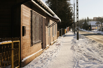 Obraz premium Snowy residential street with sidewalk and houses. Winter neighborhood scenery with icy pavement and sunlight. Quiet suburban area during cold weather. Urban daily life in winter season.