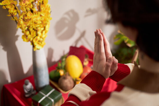 Young Asian woman clasping hands in prayer gesture in front of traditional holiday altar with fruit and yellow flowers, celebrating Tet holiday with respectful posture