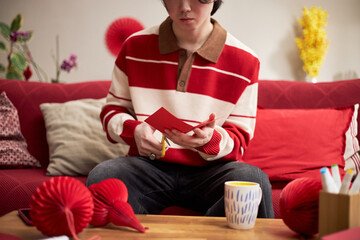 Asian teenager boy sitting on sofa cutting red paper with scissors preparing holiday decorations for Tet celebration surrounded by festive ornaments and craft supplies