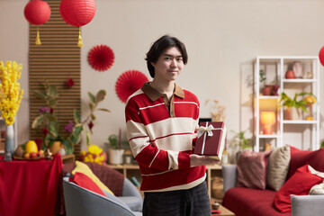 Naklejka premium Portrait of young adult Asian man smiling and holding gift box, while standing in decorated living room with Tet holiday ornaments and festive plants visible in background